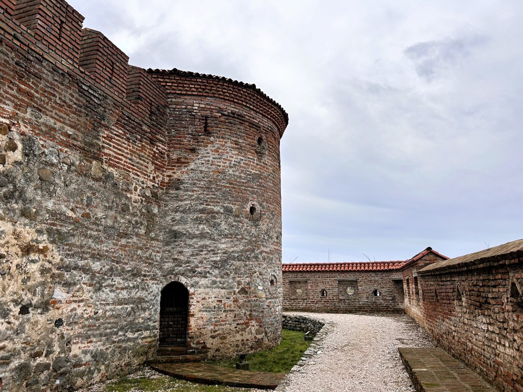 A stone fortress wall with a circular tower, featuring a small entrance and a pathway leading into the structure. The sky is overcast, adding to the historic atmosphere.