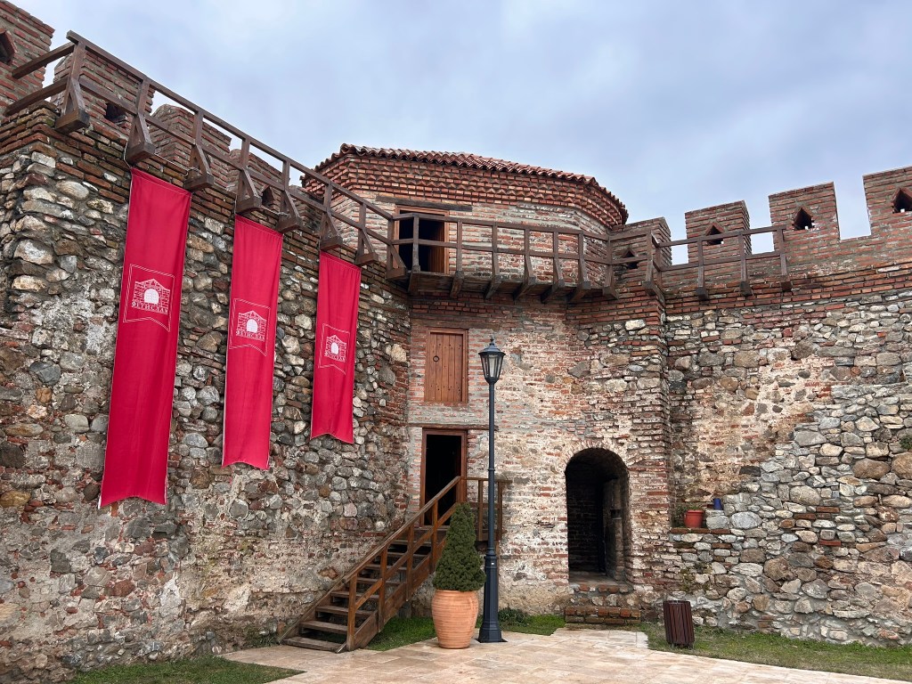 Exterior view of a historic stone castle with red banners, wooden balconies, and a cobblestone path.