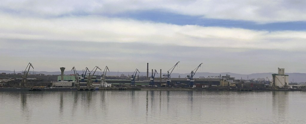 A panoramic view of a port with numerous cranes and industrial buildings along the water's edge, reflecting on the calm surface.