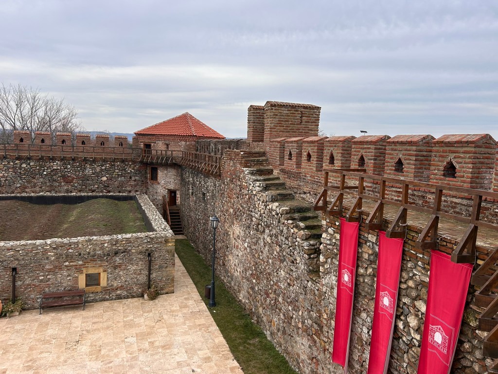 View of a historic stone fortress with a courtyard, featuring red banners and a brick tower under a cloudy sky.