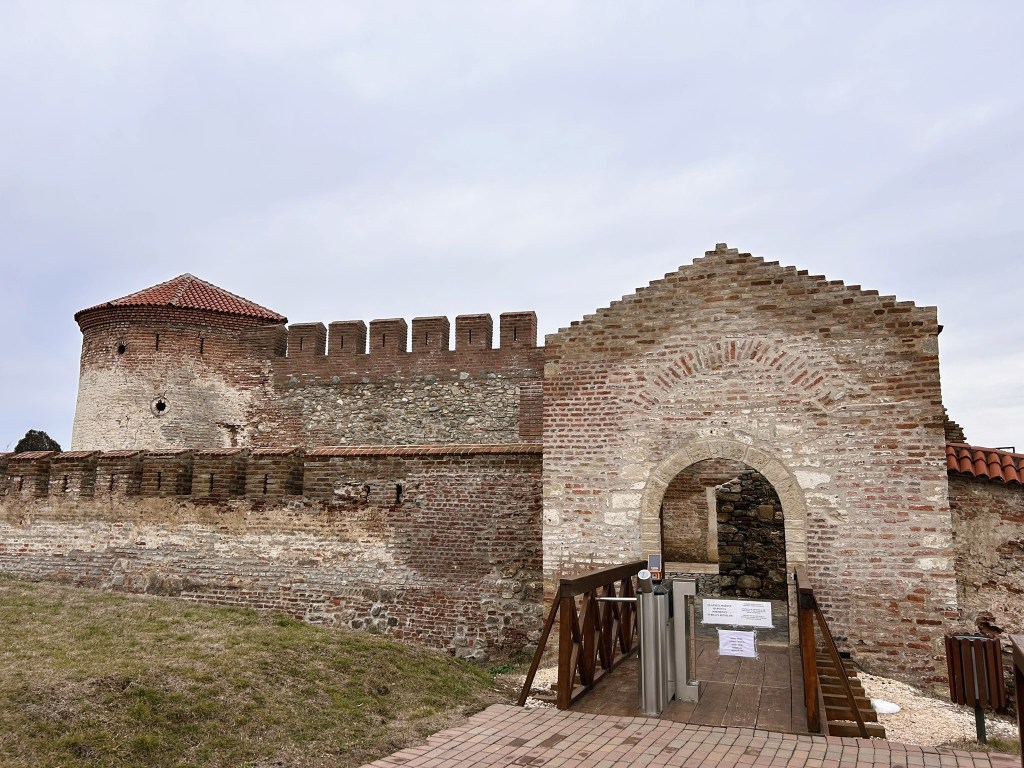 A historic stone fortress featuring a round tower on the left, a brick wall with battlements, and an arched entrance surrounded by a wooden walkway.