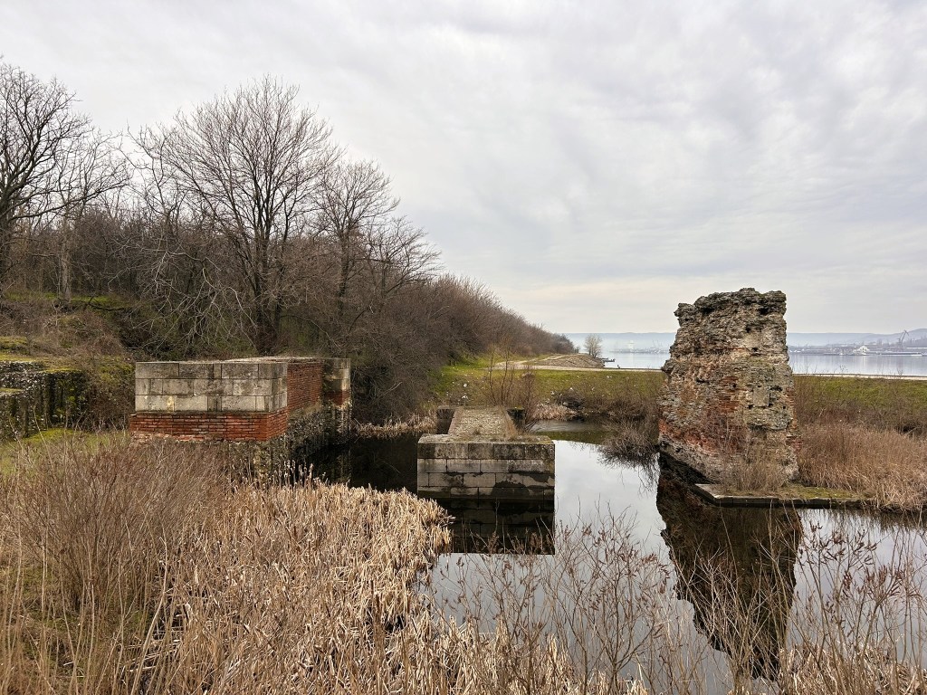 Ruins of brick structures near a calm waterbody, surrounded by dry reeds and trees, under a cloudy sky.