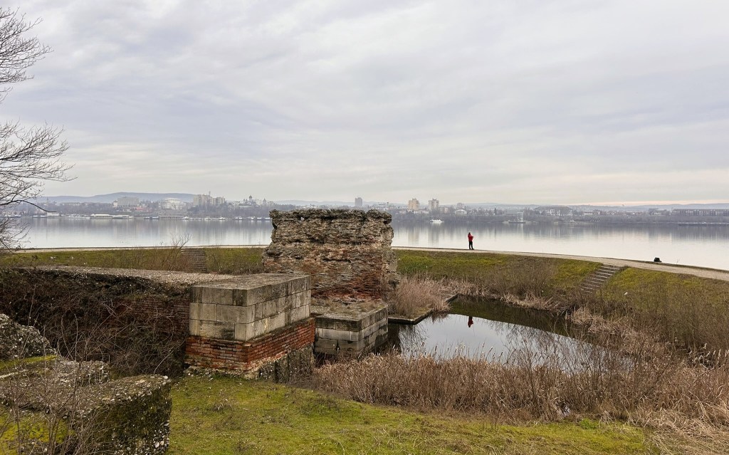 A partially ruined structure with brick and stone walls near a calm water body, reflecting the skyline in the background, with a person standing on the shore in the distance.