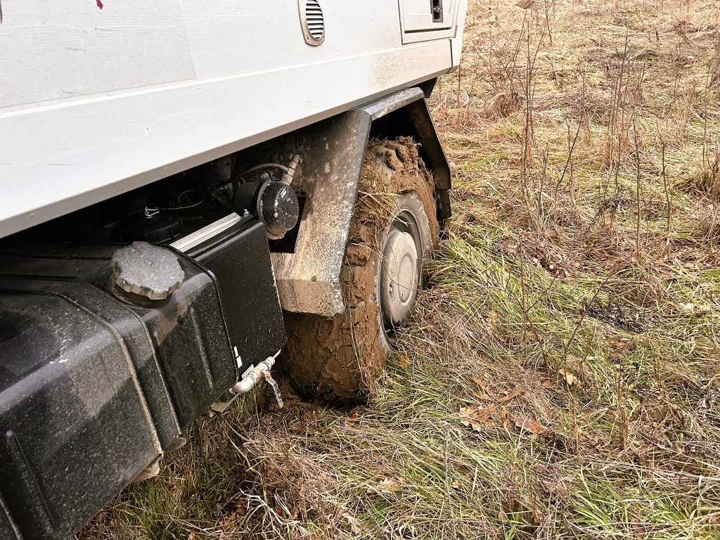 Close-up of a muddy tyre of a vehicle stuck in the grass, showing dirt on the rim and surrounding vegetation.