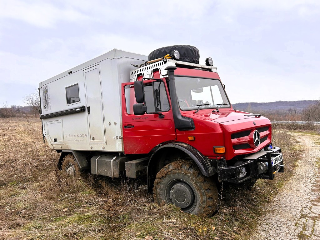 A red off-road vehicle with a grey camper body is partially stuck in a muddy field, surrounded by sparse vegetation and hills in the background.