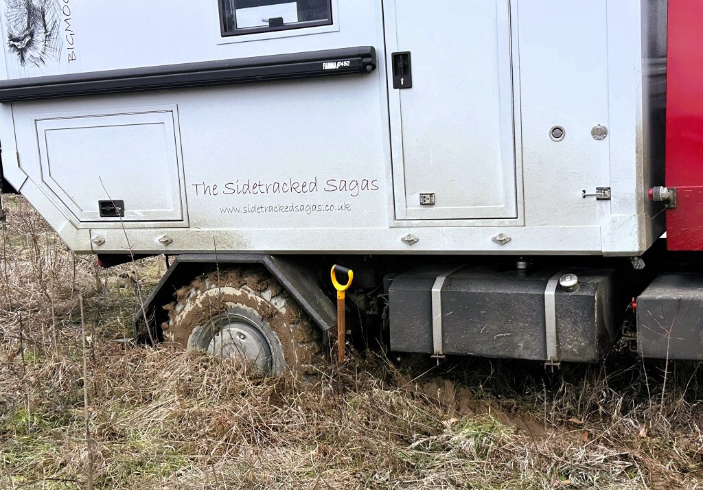 A partially buried off-road vehicle with muddy wheels, set in a grassy area with some overgrown vegetation.