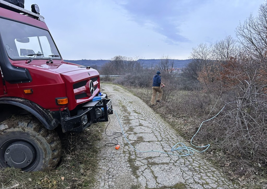 A red Mercedes off-road vehicle parked on a gravel path with a person in a blue jacket walking nearby, surrounded by bare trees and a cloudy sky.