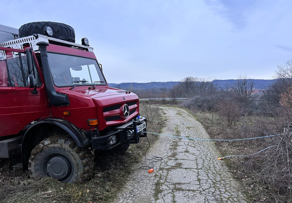 A red Mercedes off-road vehicle parked on a rugged dirt track, with a winch cable attached, under a cloudy sky.