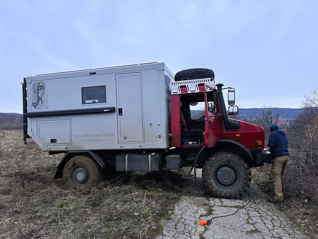 A red 4x4 vehicle with a grey camper shell parked on a rural road, with a person in a blue hoodie attending to the vehicle. The landscape is grassy with some shrubs in the background.