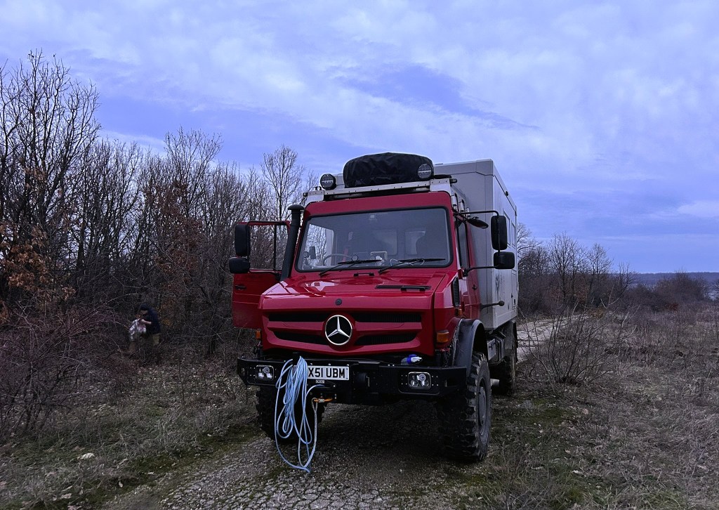 A red Mercedes off-road vehicle parked on a gravel path surrounded by sparse trees and bushes under a cloudy sky.