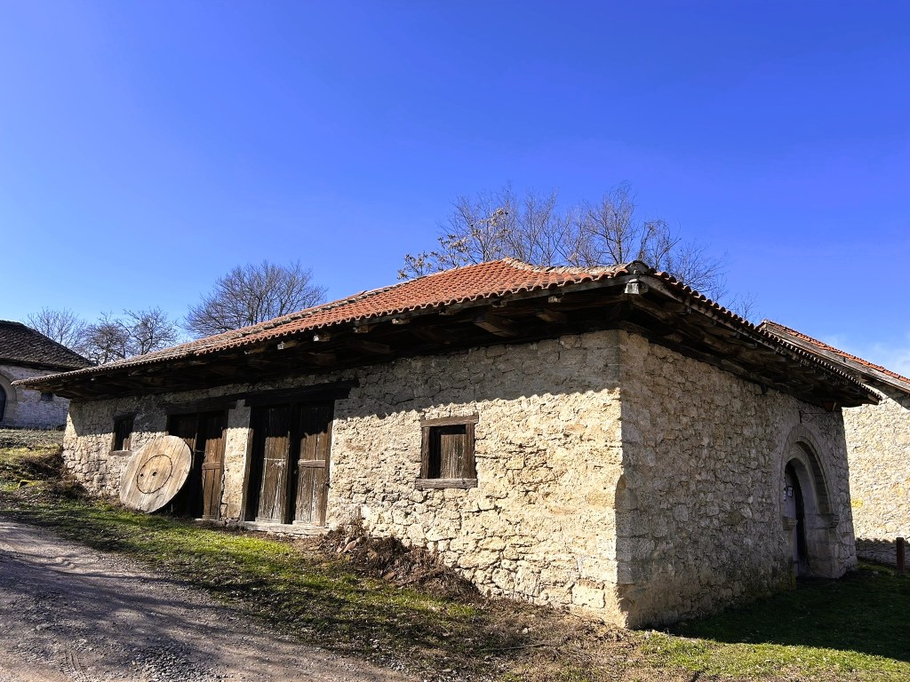 A rustic stone building with a tiled roof, featuring wooden doors and a circular wooden shield leaning against the wall, set against a clear blue sky and grassy ground.