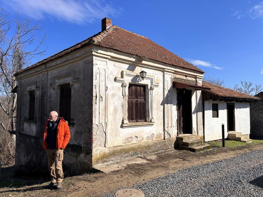 A weathered, traditional house with ornate details and wooden windows, featuring a man in an orange jacket standing in front.
