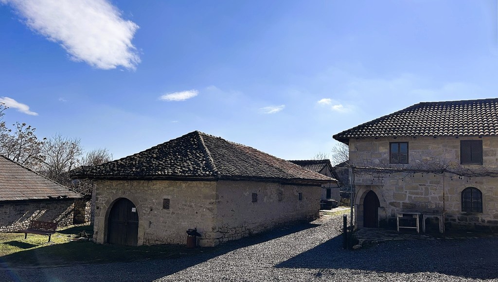 A view of traditional stone buildings with sloped roofs under a clear blue sky, surrounded by gravel pathways and sparse trees.