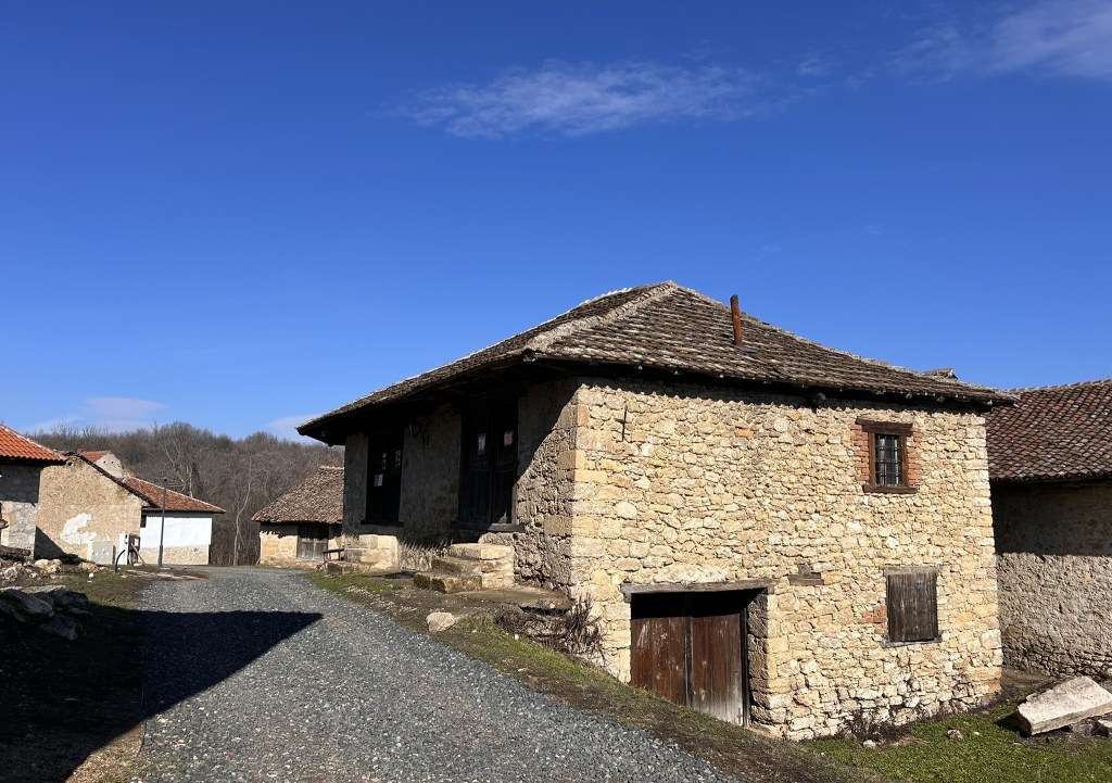 A traditional stone house with a sloped roof, located on a gravel pathway, surrounded by similar rustic buildings under a clear blue sky.
