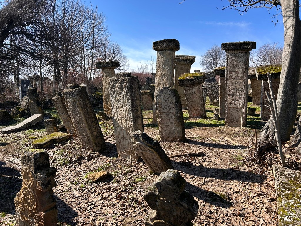 A scene of an ancient graveyard with several upright and leaning tombstones, some adorned with carvings and moss, surrounded by bare trees under a clear blue sky.