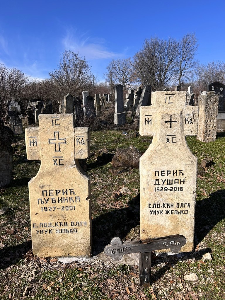 Two weathered gravestones in a cemetery, one featuring the name Перик Пубинка (1927-2001) and the other Перик Душан (1928-2016), alongside a wooden cross marked 'Душан'. The background includes various other gravestones and sparse vegetation under a clear blue sky.