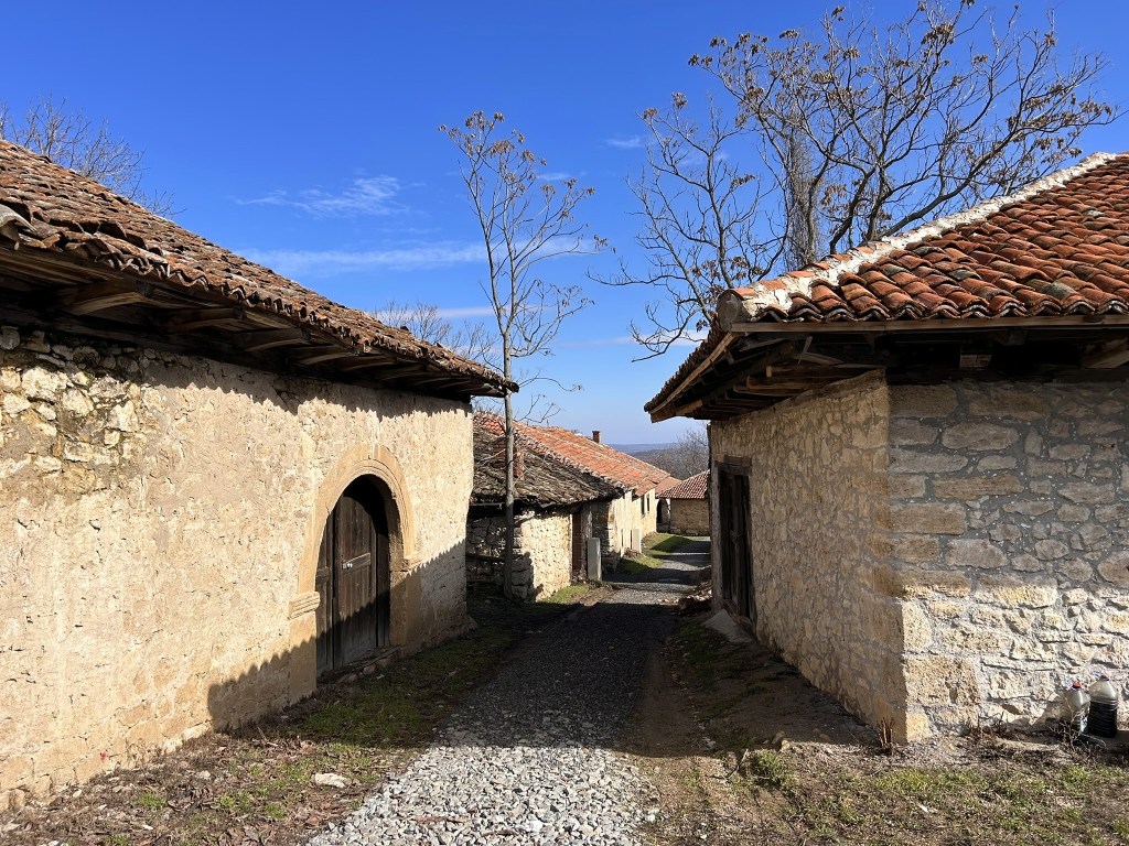 A narrow pathway between rustic stone houses with tiled roofs under a clear blue sky. Trees are visible in the background, suggesting a serene rural setting.