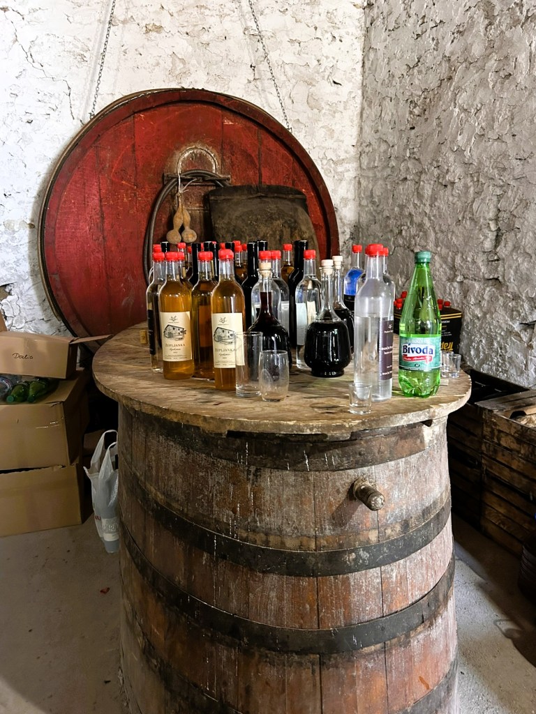 A rustic wooden barrel serving as a table, topped with various bottles of spirits and a sparkling water bottle, set against a textured stone wall.