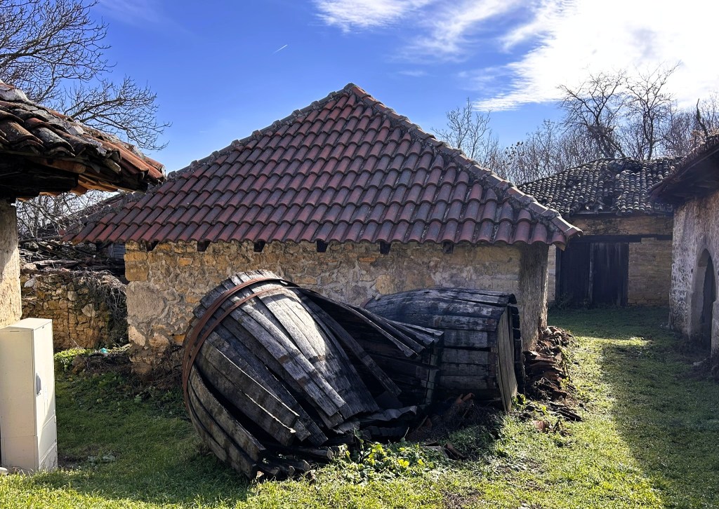An old stone building with a tiled roof beside two large, tipped wooden barrels in a grassy area, with clear blue skies and bare trees in the background.