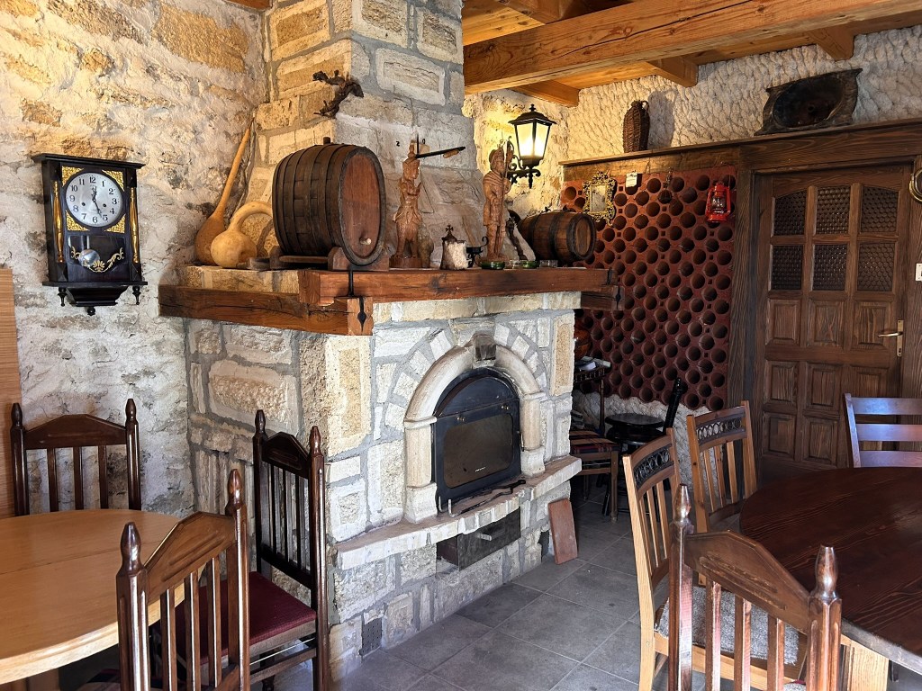 A rustic dining area featuring a stone fireplace, wooden furniture, an antique clock, and decorative items including barrels and carvings.
