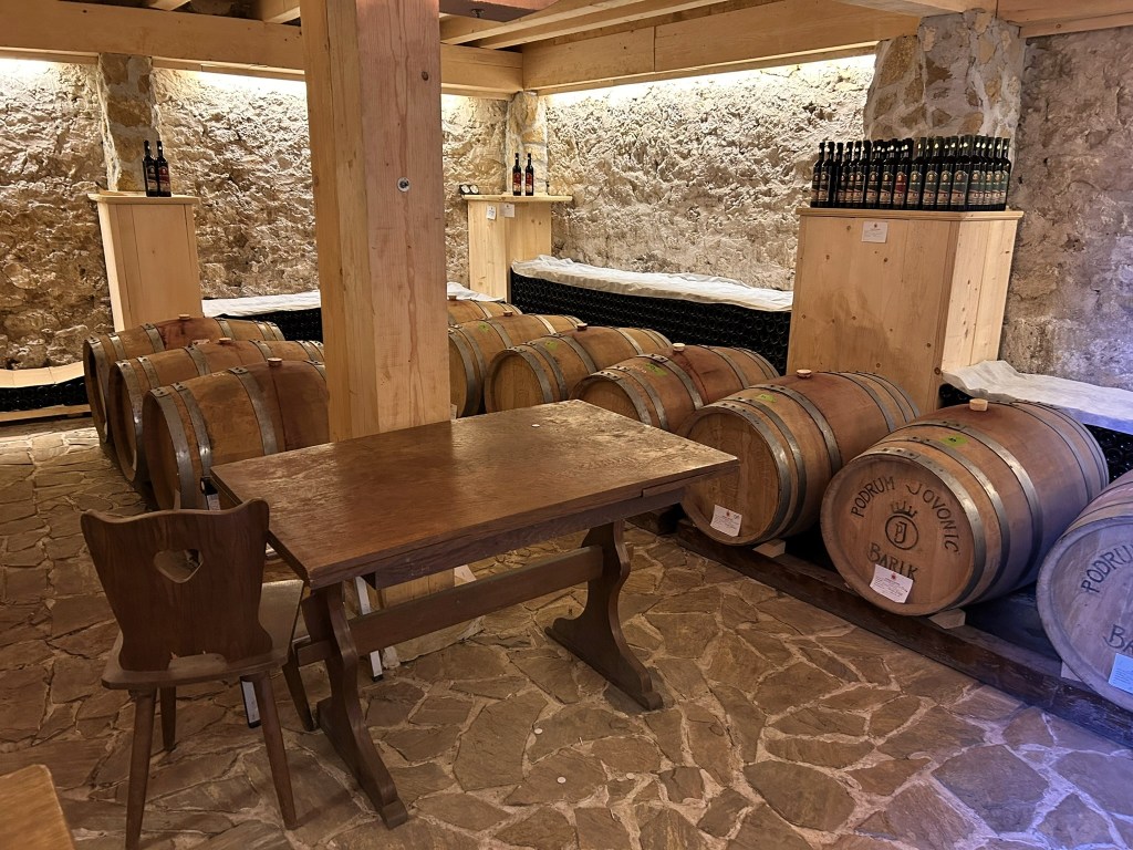 Interior of a wine cellar featuring wooden barrels, a wooden table with a chair, and stone walls.