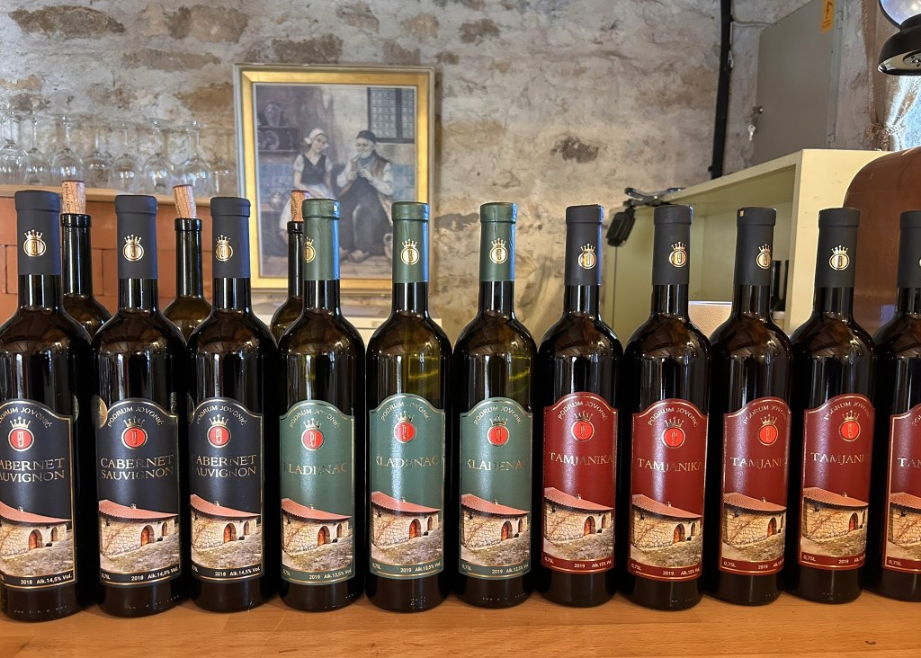A selection of wine bottles arranged in a row, featuring various labels such as Cabernet Sauvignon, Kladihnac, and Tamjanika, set against a rustic background.
