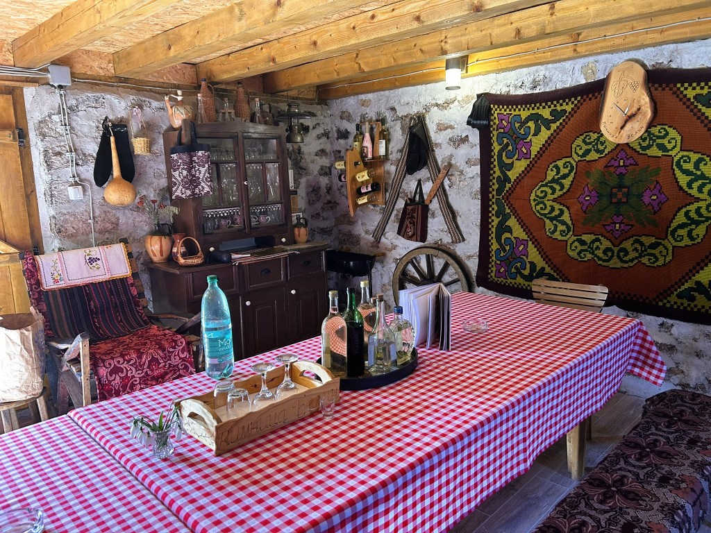 A rustic interior featuring a wooden ceiling, stone walls, and a spacious table covered with a red and white checkered tablecloth. The room contains traditional decor, including woven tapestries, glass bottles, and wooden furniture, creating a warm, homey atmosphere.