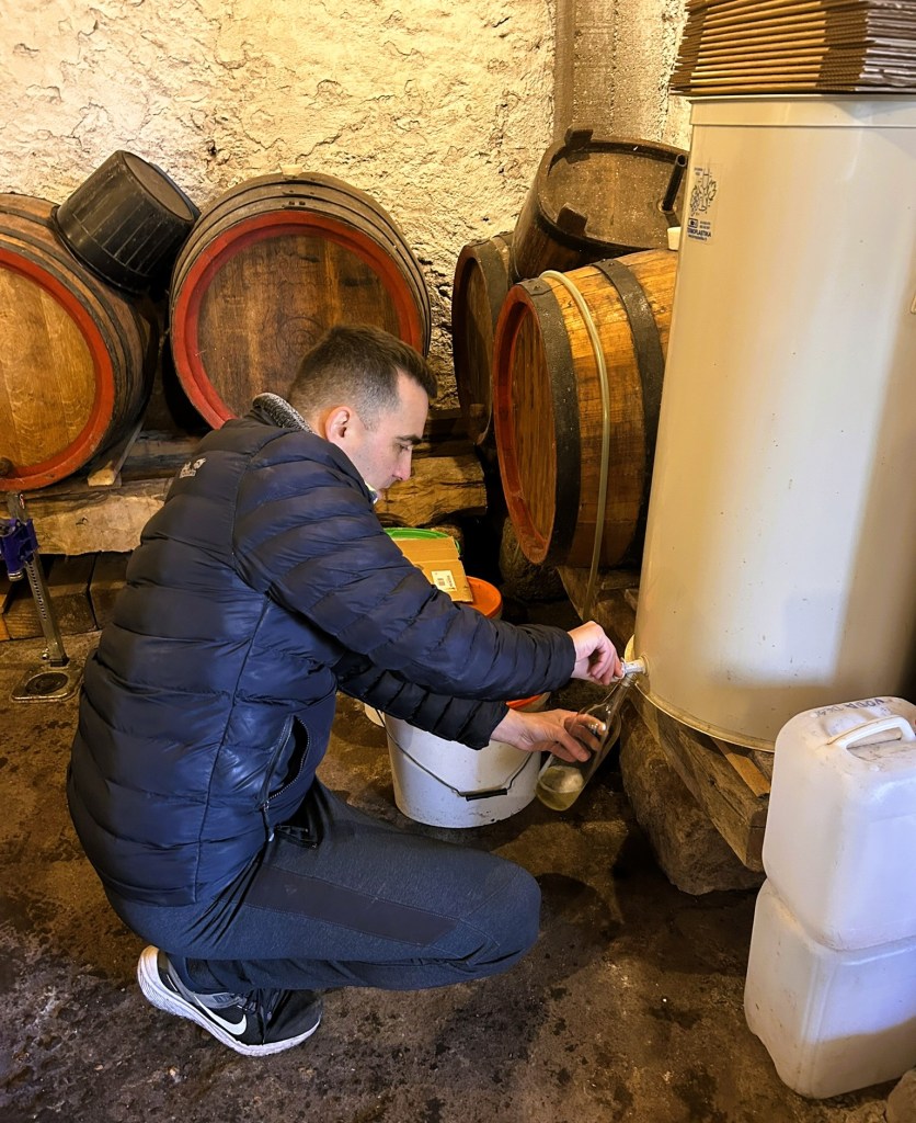 A person in a puffer jacket is crouched down, filling a glass bottle from a large container in a rustic cellar surrounded by wooden barrels.