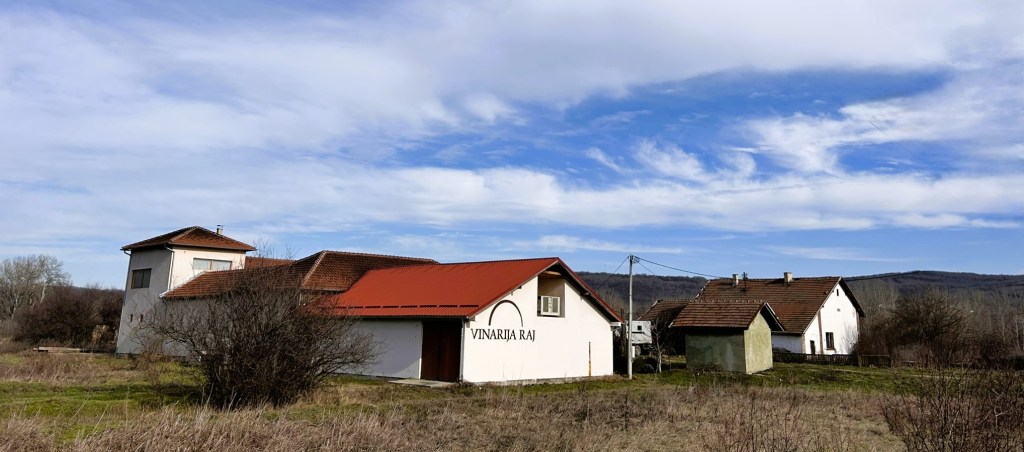 A rural scene featuring several houses with tiled roofs, surrounded by overgrown grass and trees, under a partly cloudy sky.