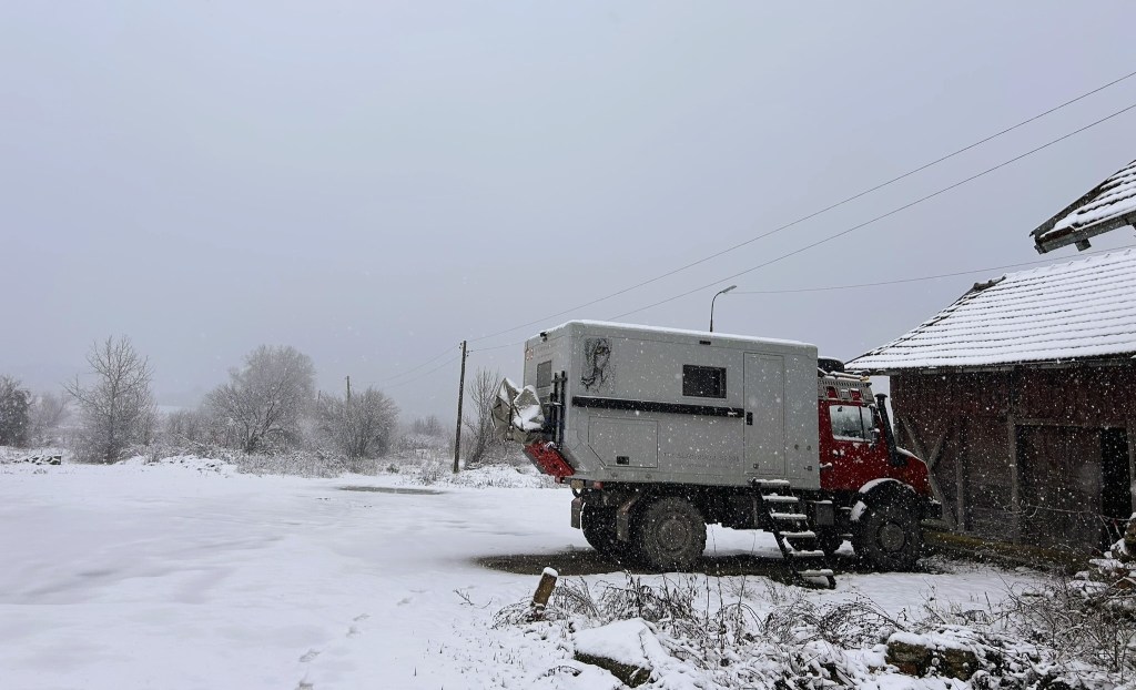 A red and grey overland vehicle parked in a snowy landscape near a wooden barn, with trees in the background and falling snow.