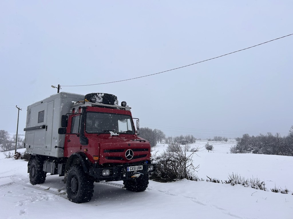 A red Mercedes truck with an adventure camper mounted on the back, parked in a snowy landscape with falling snow and distant trees.