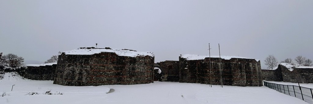 A panoramic view of an ancient stone fortress partially covered in snow, under a grey sky.