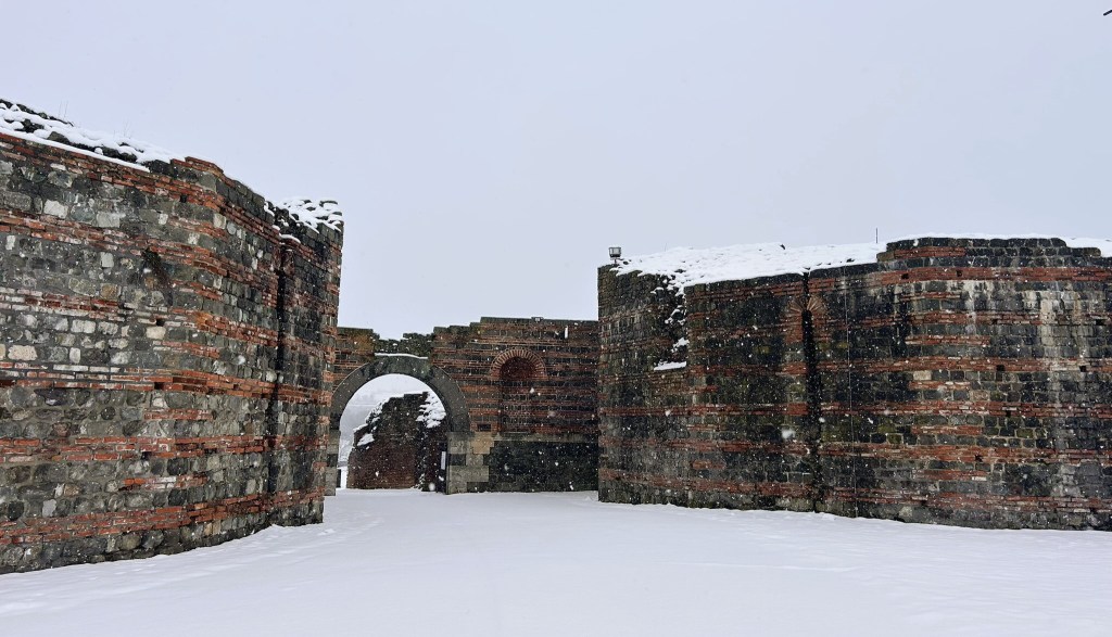 Snow-covered ruins of ancient brick walls with a prominent archway, set against a grey sky.