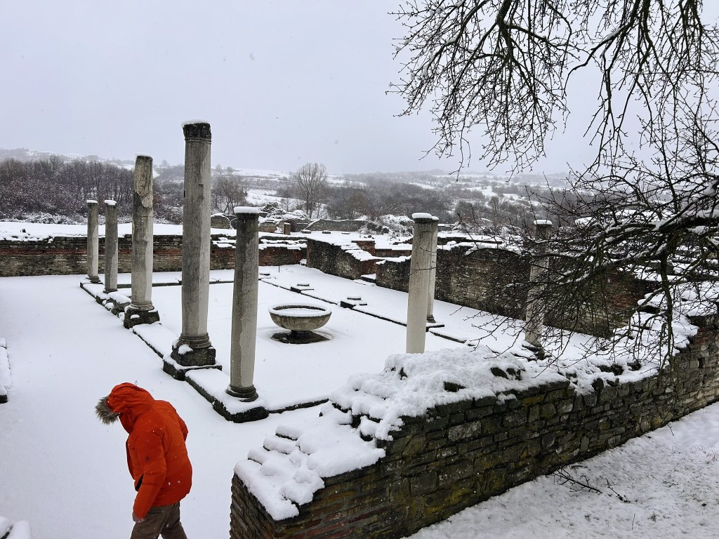 A person in an orange coat walking through snow-covered ancient ruins, featuring columns and remnants of stone structures, under a grey, snowy sky.