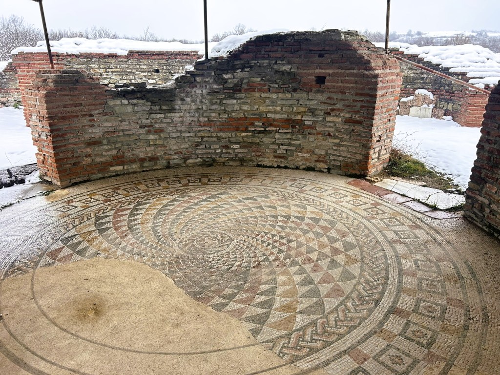 A circular mosaic with intricate geometric patterns in muted colours, surrounded by brick and stone walls, with snow visible outside.