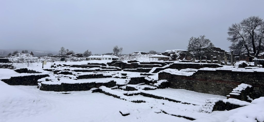 Snow-covered archaeological ruins with stone foundations and structures under a cloudy sky.