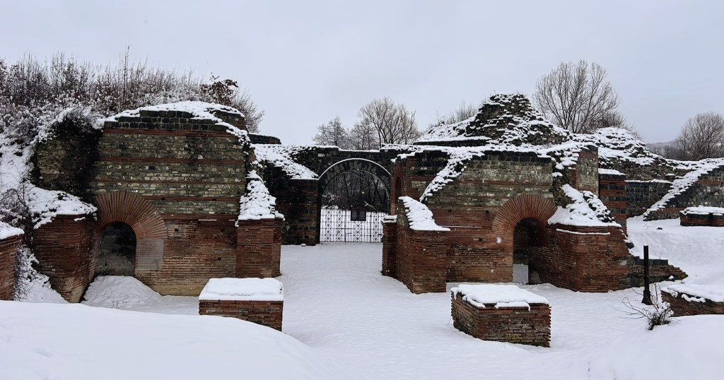 Snow-covered ruins of an ancient structure, featuring brick arches and a wrought iron gate in a wintry landscape.