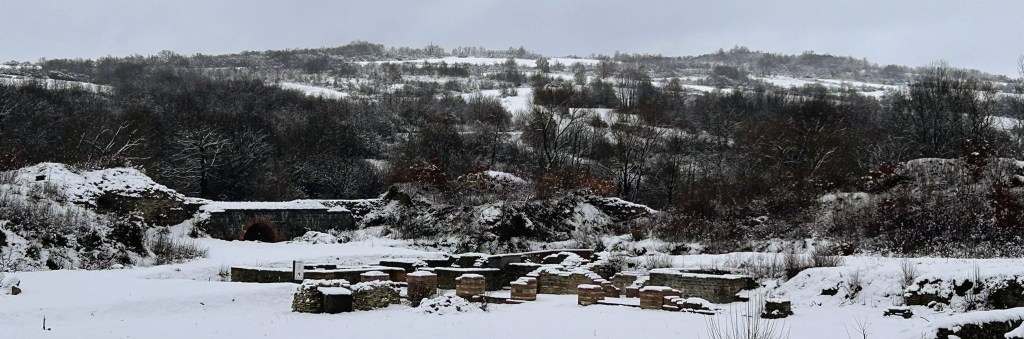 A snowy landscape featuring archaeological ruins and rolling hills in the background, with trees partially covered in snow.