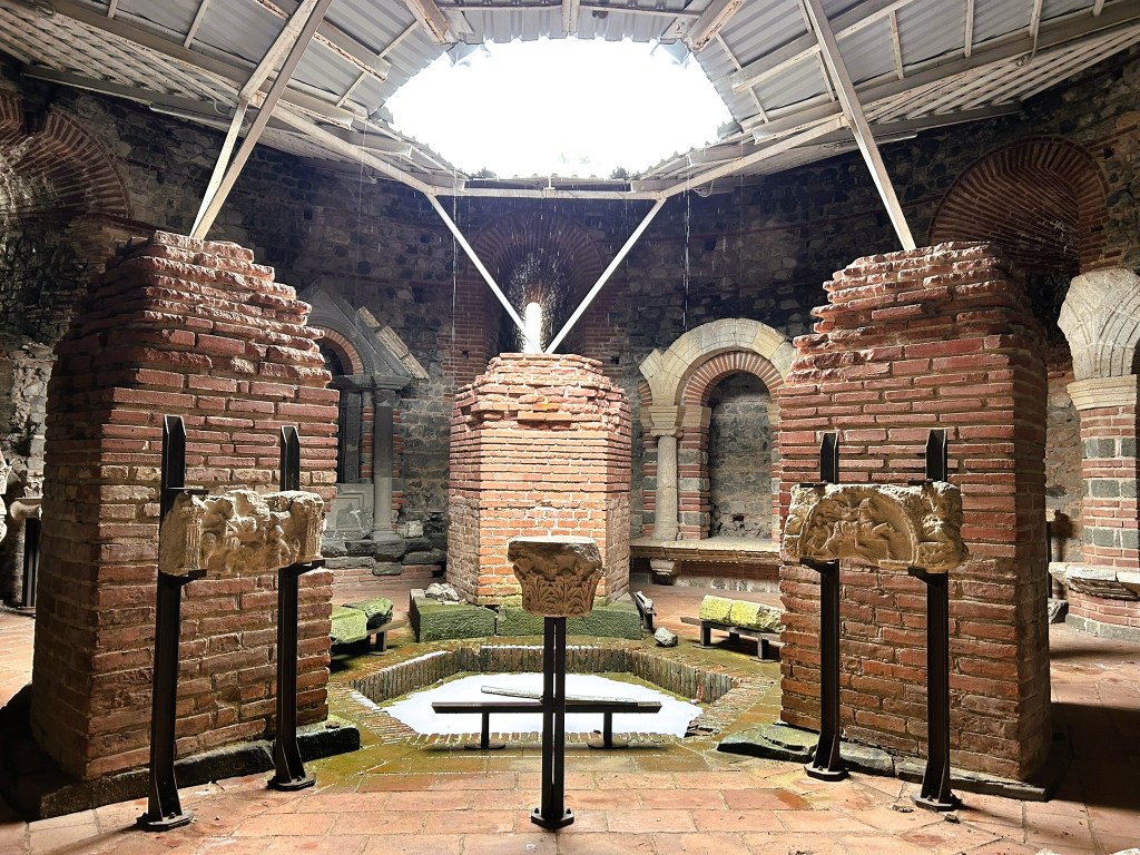 Interior view of an ancient building featuring brick columns, stone carvings on metal stands, and a circular water feature beneath a skylight.