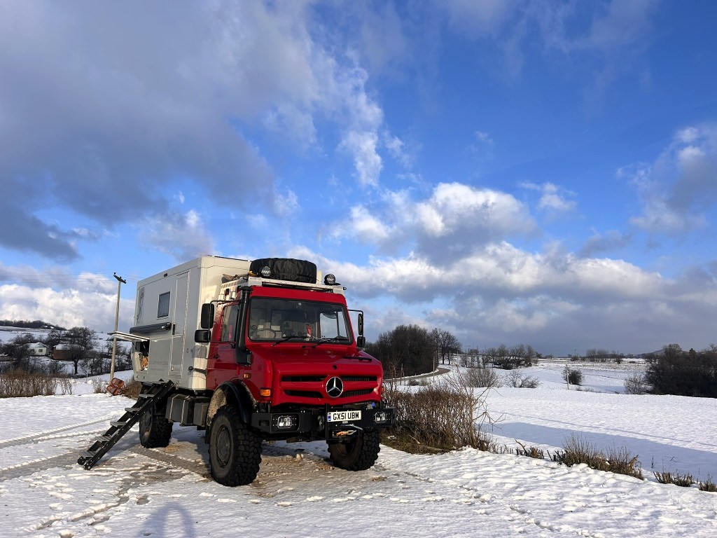 A red Mercedes truck with an attached camper setup parked in a snowy landscape under a blue sky with clouds.
