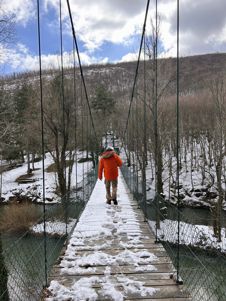 A person wearing an orange jacket walking across a snowy suspension bridge surrounded by trees and a river.