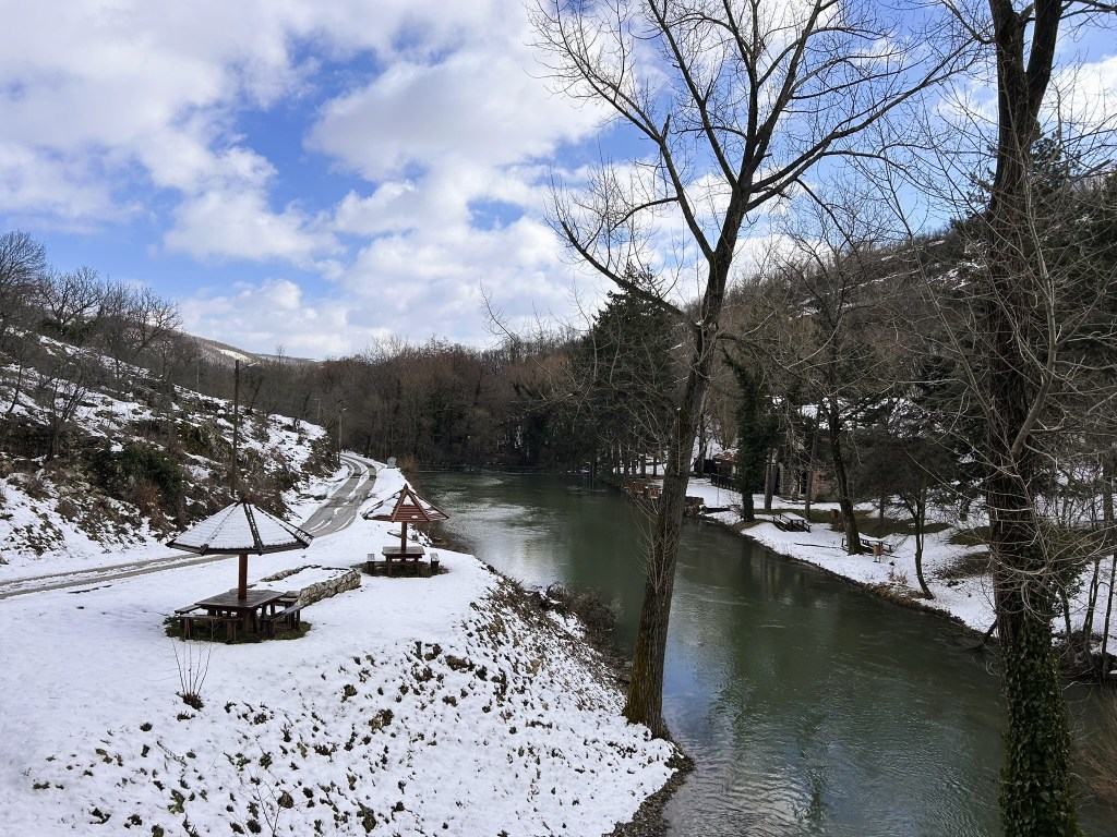 A serene landscape featuring a river bordered by snow-covered banks, with wooden gazebos and benches on the shore, framed by leafless trees and a partly cloudy sky.