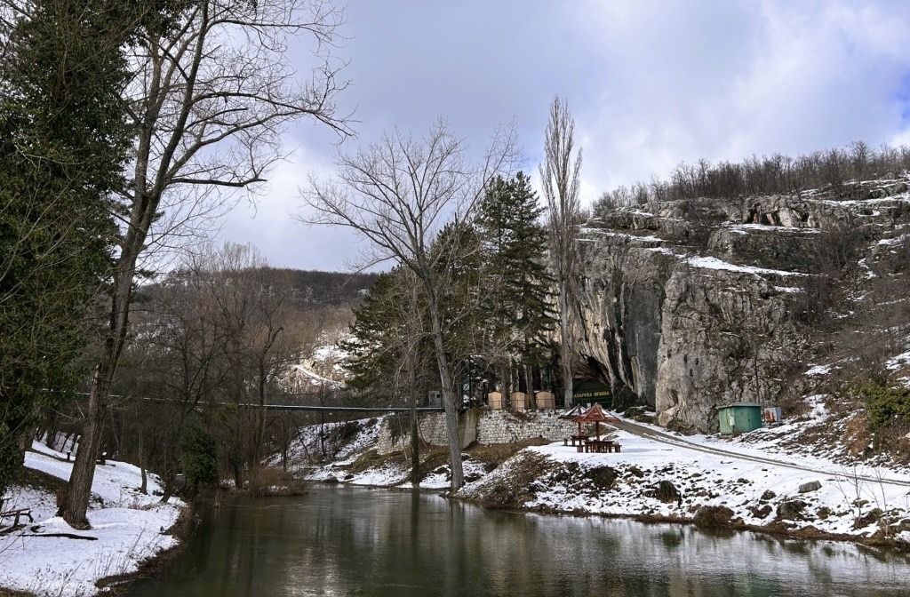 A serene winter landscape featuring a calm river, surrounded by trees and rocky cliffs. Snow covers the ground, while a small wooden structure and a green shed are visible along the riverbank.