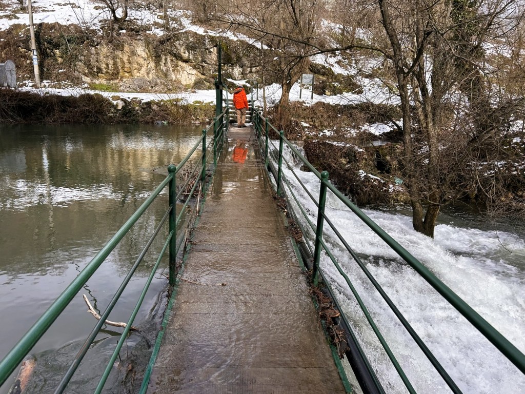 A person in an orange coat walking on a narrow concrete bridge surrounded by flowing water, with snow on the ground and trees in the background.