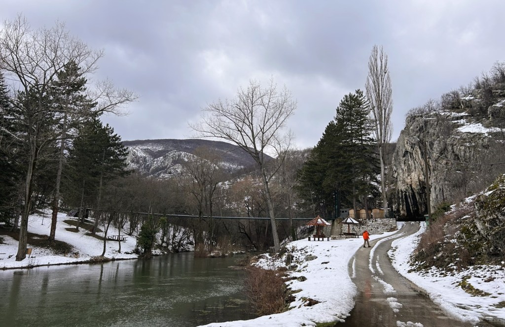 A scenic view of a river bordered by snow-covered ground and trees, with a person walking along a path. In the background, hills rise under a cloudy sky.