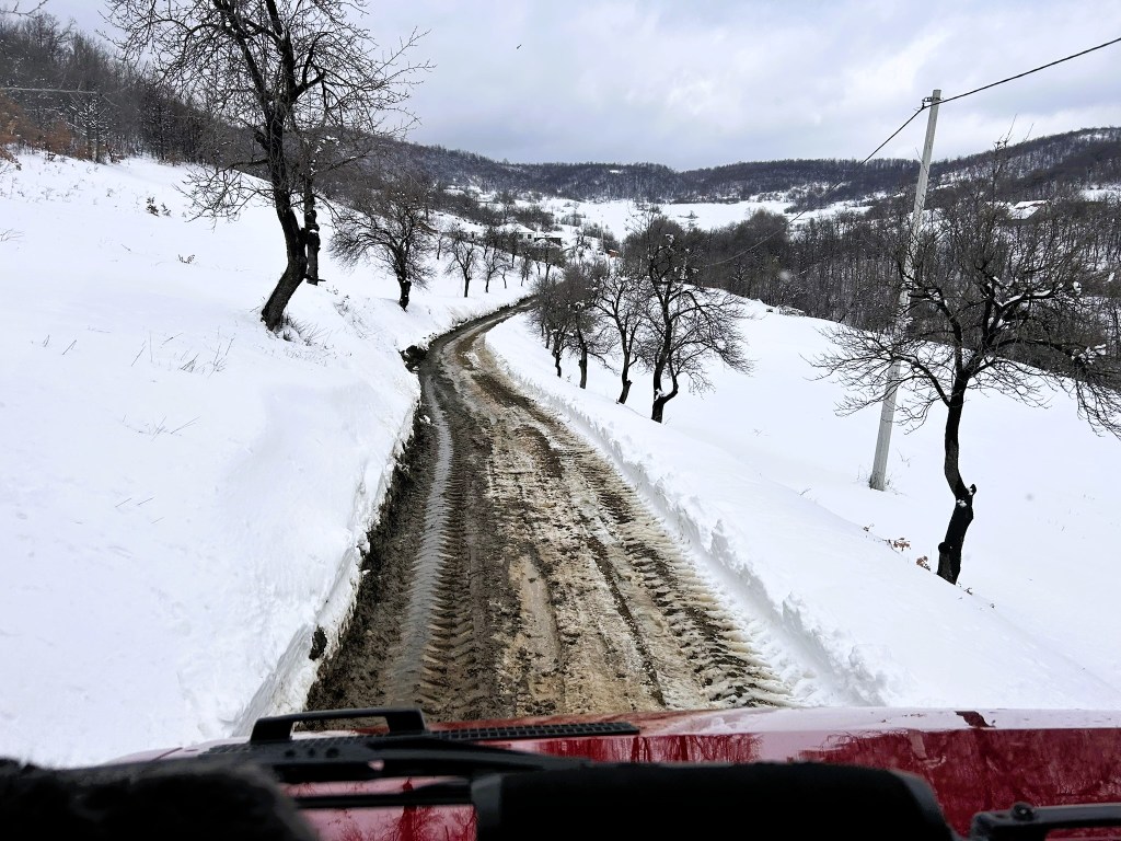 A snowy road winding through a winter landscape, flanked by bare trees, with visible tire tracks in the mud.