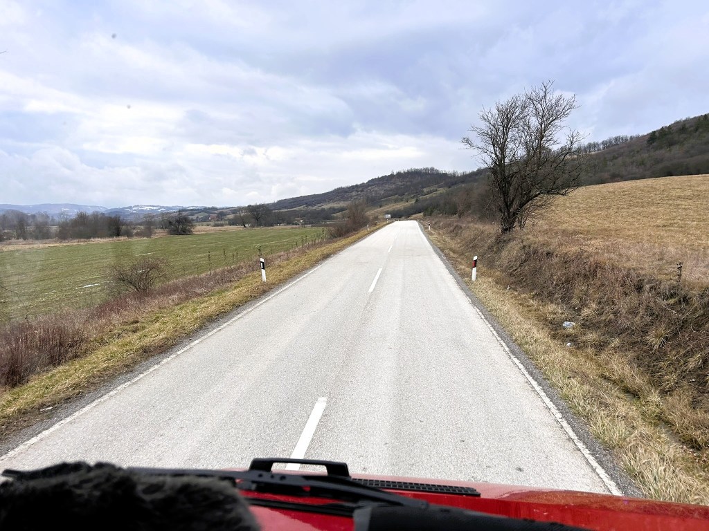 A straight road lined with grassy fields and a few trees, under a cloudy sky.
