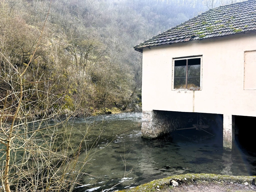 A partially submerged building beside a river, surrounded by bare trees and a misty hillside.
