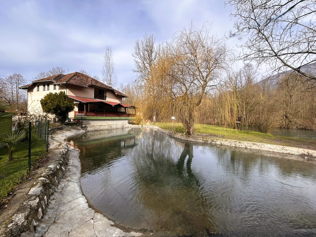 A tranquil scene featuring a house beside a calm pond, with trees and a grassy area in the background.