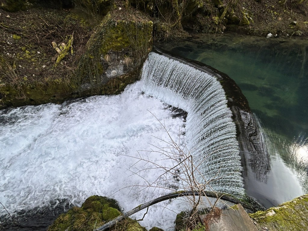 Aerial view of a waterfall cascading over a rocky ledge into a clear pool of water, surrounded by lush greenery.